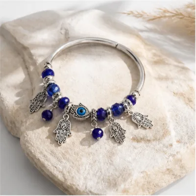 Pulseira prateada com olho azul e mãos da sorte sobre pedra clara