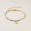 Pulseira dourada com missangas multicolores, elos ovais e pendente de trevo sobre fundo claro