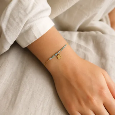 Pulseira dourada com missangas azul-claro e turquesa, elos ovais e pendente de trevo apresentada no pulso