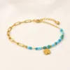 Pulseira dourada com missangas azul-claro e turquesa, elos ovais e pendente de trevo sobre fundo claro