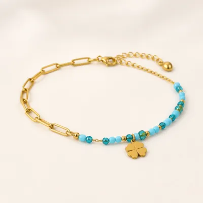 Pulseira dourada com missangas azul-claro e turquesa, elos ovais e pendente de trevo sobre fundo claro